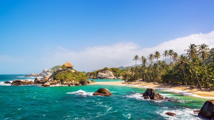Plage du parc national Tayrona bordée de cocotiers, avec un abri au toit de paille sur un promontoire rocheux et une mer turquoise