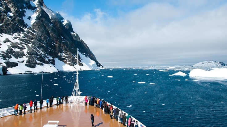 Passagers à la proue d&rsquo;un navire d&rsquo;expédition naviguant entre des blocs de glace, face à une côte montagneuse enneigée de l&rsquo;Antarctique